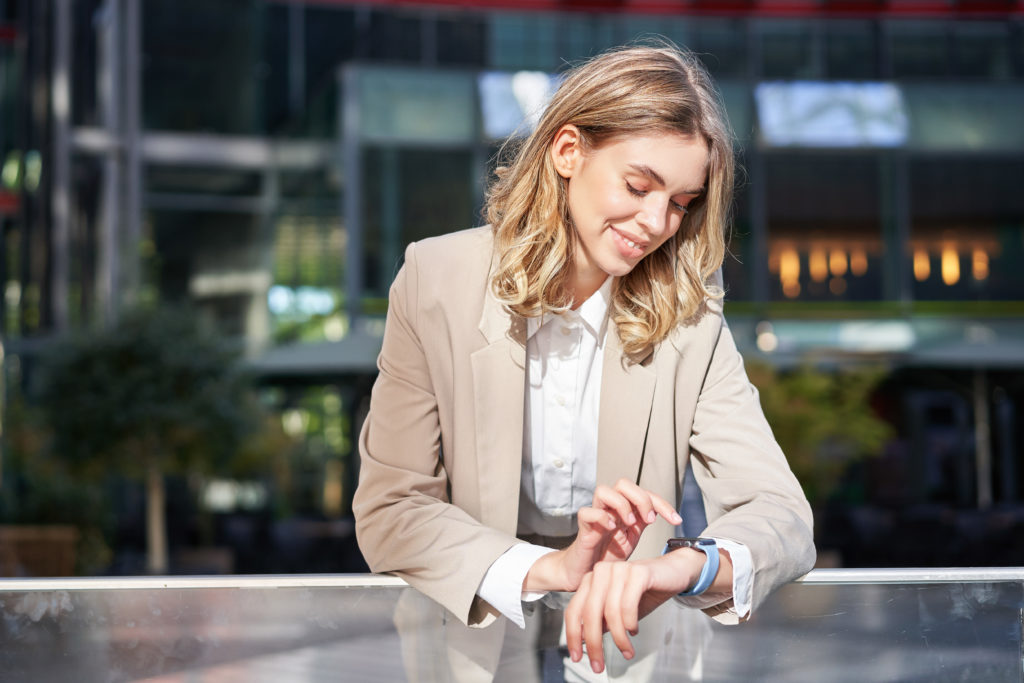 Une femme d’affaires souriante regarde sa montre numérique, lit des messages ou vérifie l’heure, se tient dehors en costume de bureau beige. Elle semble heureuse dans son travail mais se tient prête pour de nouvelles opportunités professionnelles.